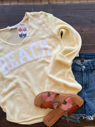 Beige 'BEACH' shirt with earrings, sandals, and denim shorts on a wooden surface