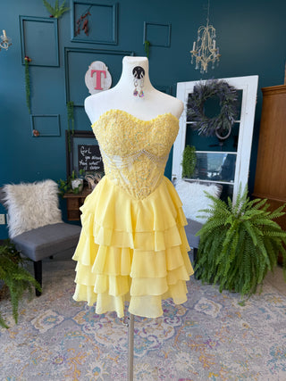 A yellow strapless sweetheart dress with a beaded corset-style bodice and tiered ruffle skirt displayed on a white mannequin in a decorated room with teal walls, plants, and a patterned rug.