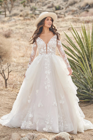 Woman in a white wedding dress with long sleeves and a hat standing in a desert landscape.
