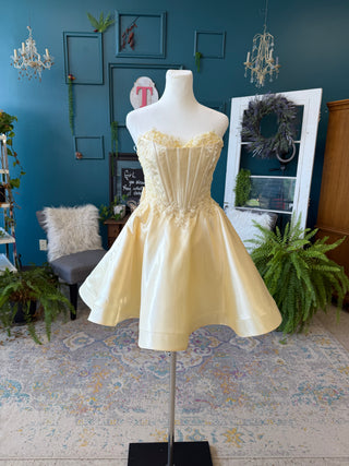 A pale yellow strapless satin mini dress with a corset-style bodice and flared skirt displayed on a white mannequin in a styled room with blue walls, plants, and decorative furniture.