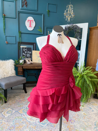 A bright pinkish-red tiered halter dress displayed on a mannequin torso in a styled room with teal walls, plants, and furniture.