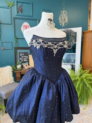 A navy blue off-the-shoulder quinceañera dress with silver rhinestone embellishments and a flared skirt, displayed on a white mannequin in a decorated boutique setting.