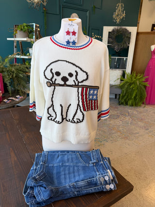 White sweater with a dog design and American flag on a mannequin, with blue jeans folded below, in an indoor setting.