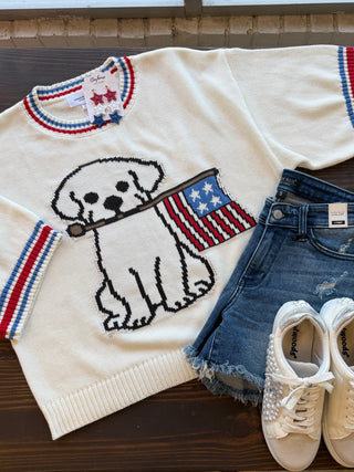 White sweater with dog and flag design, denim shorts, and white sneakers on a wooden surface.
