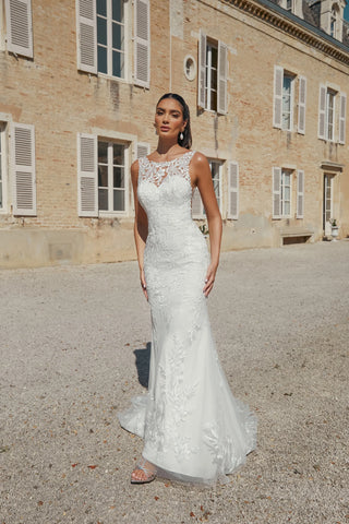 Woman in a white lace wedding dress standing in front of a building with shutters.
